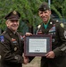 Two U.S. Army soldiers pose together holding a framed Certificate of Appreciation during what appears to be a military retirement or awards ceremony. The officer on the left, wearing two-star Major General rank insignia and a 10th Mountain Division patch, is presenting the certificate to a Master Sergeant on the right who is wearing the Medal of Honor around his neck. The certificate is addressed to Master Sergeant Caleb Barrieau, United States Army.