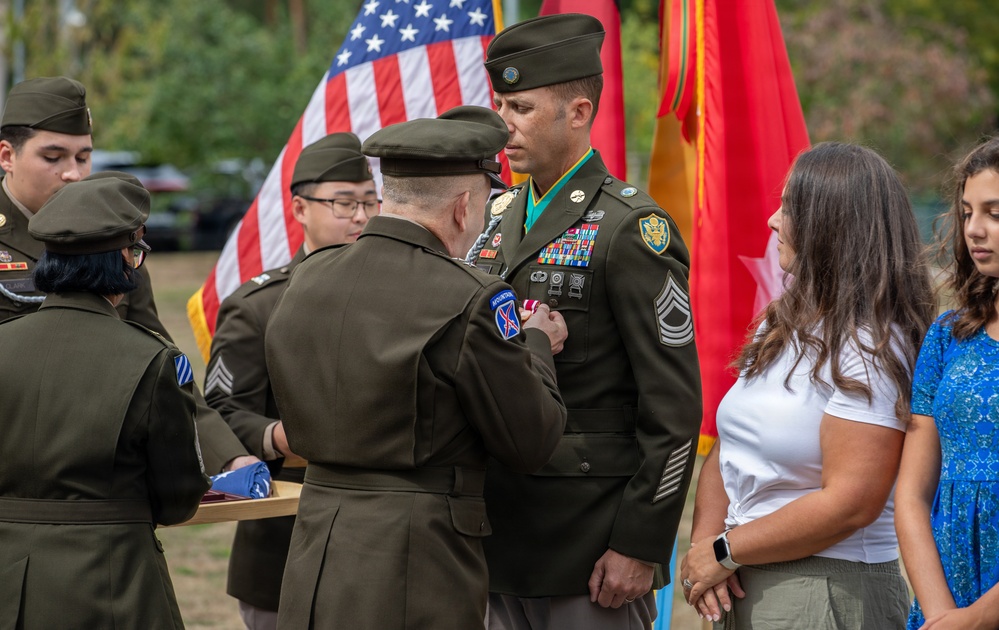 A U.S. Army military ceremony is taking place outdoors, where a senior officer is pinning a medal or award onto a Master Sergeant or Sergeant First Class in dress green uniform. The recipient is wearing multiple service ribbons, a green beret-style flash on his shoulder patch, and a Special Forces tab, suggesting this may be a decoration or retirement ceremony. Civilians, likely family members, stand nearby alongside other uniformed soldiers holding a folded flag.