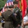 A U.S. Army military ceremony is taking place outdoors, where a senior officer is pinning a medal or award onto a Master Sergeant or Sergeant First Class in dress green uniform. The recipient is wearing multiple service ribbons, a green beret-style flash on his shoulder patch, and a Special Forces tab, suggesting this may be a decoration or retirement ceremony. Civilians, likely family members, stand nearby alongside other uniformed soldiers holding a folded flag.
