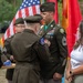 A U.S. Army military ceremony is taking place outdoors, where a senior officer is pinning a medal or award onto a Master Sergeant or Sergeant First Class in dress green uniform. The recipient is wearing multiple service ribbons, a green beret-style flash on his shoulder patch, and a Special Forces tab, suggesting this may be a decoration or retirement ceremony. Civilians, likely family members, stand nearby alongside other uniformed soldiers holding a folded flag.