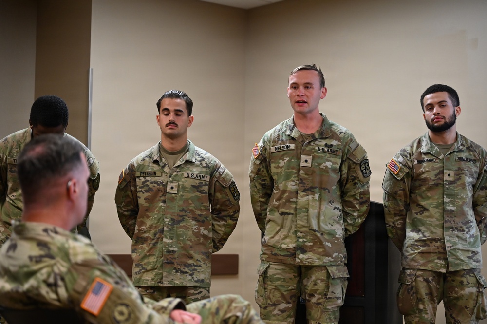 A group of U.S. Army soldiers in OCP (Operational Camouflage Pattern) uniforms are standing at attention or parade rest in what appears to be an indoor briefing or formation setting. Visible name tapes identify at least three soldiers: PETERLIN, NELSON, and a partially visible name ending in '-AMMAD'. One soldier appears to be speaking or addressing someone seated in the foreground.