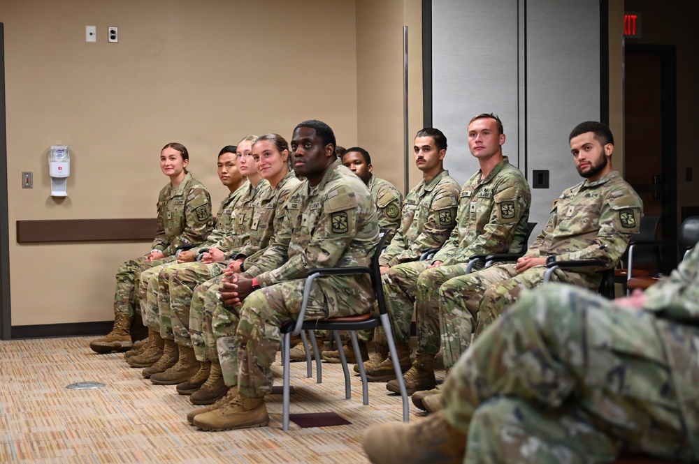 A group of approximately 10-12 U.S. Army soldiers in OCP (Operational Camouflage Pattern) uniforms are seated in rows in what appears to be a briefing room or conference facility. The soldiers are attentively facing forward, suggesting they are listening to a presentation or receiving instruction. The group is diverse in gender and ethnicity, consistent with a modern U.S. Army unit formation.