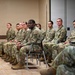 A group of approximately 10-12 U.S. Army soldiers in OCP (Operational Camouflage Pattern) uniforms are seated in rows in what appears to be a briefing room or conference facility. The soldiers are attentively facing forward, suggesting they are listening to a presentation or receiving instruction. The group is diverse in gender and ethnicity, consistent with a modern U.S. Army unit formation.