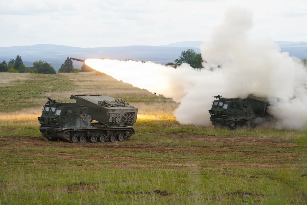 Two tracked military multiple launch rocket systems (MLRS) are positioned in an open field, with one actively firing a rocket. A large plume of white smoke and exhaust is visible from the launch, with the rocket's trail clearly visible ascending at an angle. The setting appears to be a military training exercise or live-fire demonstration in a rural, hilly landscape.