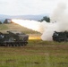 Two tracked military multiple launch rocket systems (MLRS) are positioned in an open field, with one actively firing a rocket. A large plume of white smoke and exhaust is visible from the launch, with the rocket's trail clearly visible ascending at an angle. The setting appears to be a military training exercise or live-fire demonstration in a rural, hilly landscape.