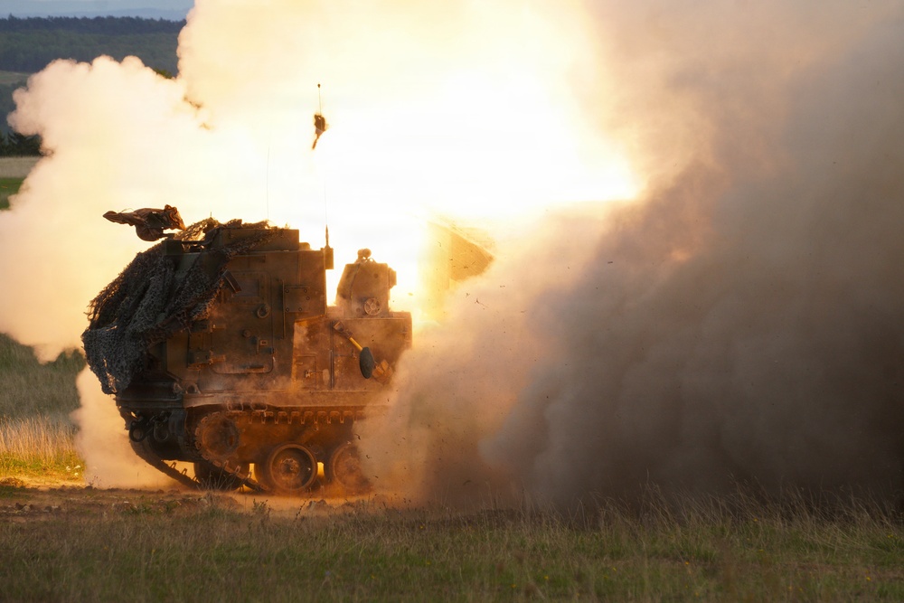 A tracked military rocket artillery system is captured mid-launch, with a rocket visibly leaving the launcher and generating a massive fireball and smoke cloud. The vehicle appears to be an M270 Multiple Launch Rocket System (MLRS) or similar platform, covered in camouflage netting. The image was taken during what appears to be a live-fire exercise or combat operation in an open field environment.