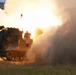 A tracked military rocket artillery system is captured mid-launch, with a rocket visibly leaving the launcher and generating a massive fireball and smoke cloud. The vehicle appears to be an M270 Multiple Launch Rocket System (MLRS) or similar platform, covered in camouflage netting. The image was taken during what appears to be a live-fire exercise or combat operation in an open field environment.