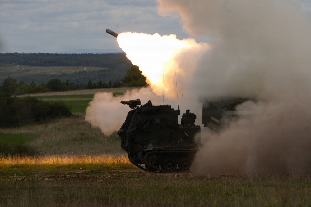 A tracked military rocket artillery system is captured mid-fire, launching a rocket with a large muzzle flash and billowing smoke cloud. The vehicle appears to be positioned in an open grassy field with a forested hillside visible in the background. The image was taken during low-light conditions, likely dusk or dawn.