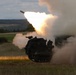 A tracked military rocket artillery system is captured mid-fire, launching a rocket with a large muzzle flash and billowing smoke cloud. The vehicle appears to be positioned in an open grassy field with a forested hillside visible in the background. The image was taken during low-light conditions, likely dusk or dawn.
