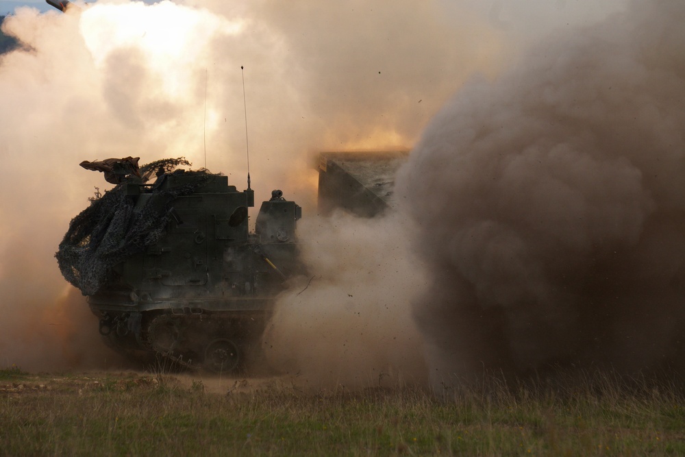 A tracked military rocket artillery system, consistent with an M270 MLRS or similar multiple launch rocket system, is captured mid-fire on an open grassy field. A massive cloud of smoke, dust, and exhaust billows around the vehicle following a rocket launch. The vehicle is draped with camouflage netting and appears to be in an active firing exercise or combat operation.