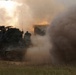 A tracked military rocket artillery system, consistent with an M270 MLRS or similar multiple launch rocket system, is captured mid-fire on an open grassy field. A massive cloud of smoke, dust, and exhaust billows around the vehicle following a rocket launch. The vehicle is draped with camouflage netting and appears to be in an active firing exercise or combat operation.