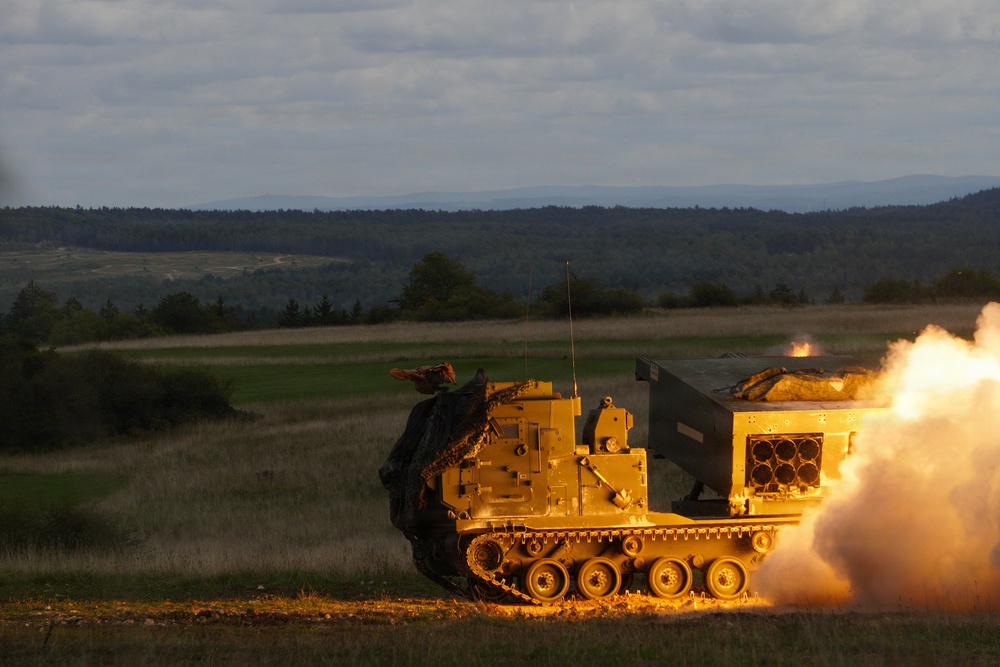 A tracked military multiple launch rocket system (MLRS) is captured mid-fire, with a rocket visibly launching from the right side of the vehicle accompanied by a large plume of smoke and flame. The vehicle appears to be an M270 MLRS or similar platform, painted in a tan/desert color scheme, positioned on an open grassy field. The image was taken during low-light conditions, likely dusk or dawn, with forested hills visible in the background.