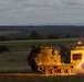 A tracked military multiple launch rocket system (MLRS) is captured mid-fire, with a rocket visibly launching from the right side of the vehicle accompanied by a large plume of smoke and flame. The vehicle appears to be an M270 MLRS or similar platform, painted in a tan/desert color scheme, positioned on an open grassy field. The image was taken during low-light conditions, likely dusk or dawn, with forested hills visible in the background.