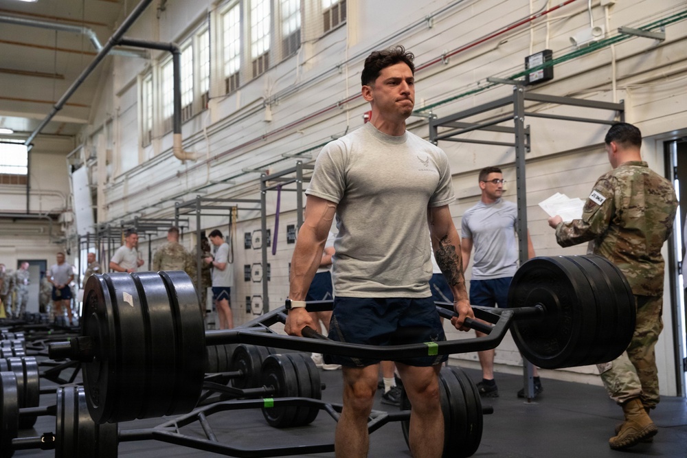 A muscular male service member in a gray U.S. Air Force t-shirt performs a trap bar (hex bar) deadlift in a military gymnasium. Multiple other personnel in PT gear and military uniforms are visible in the background, some appearing to observe or record results. The setting appears to be a formal physical fitness test or competition event.