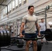 A muscular male service member in a gray U.S. Air Force t-shirt performs a trap bar (hex bar) deadlift in a military gymnasium. Multiple other personnel in PT gear and military uniforms are visible in the background, some appearing to observe or record results. The setting appears to be a formal physical fitness test or competition event.