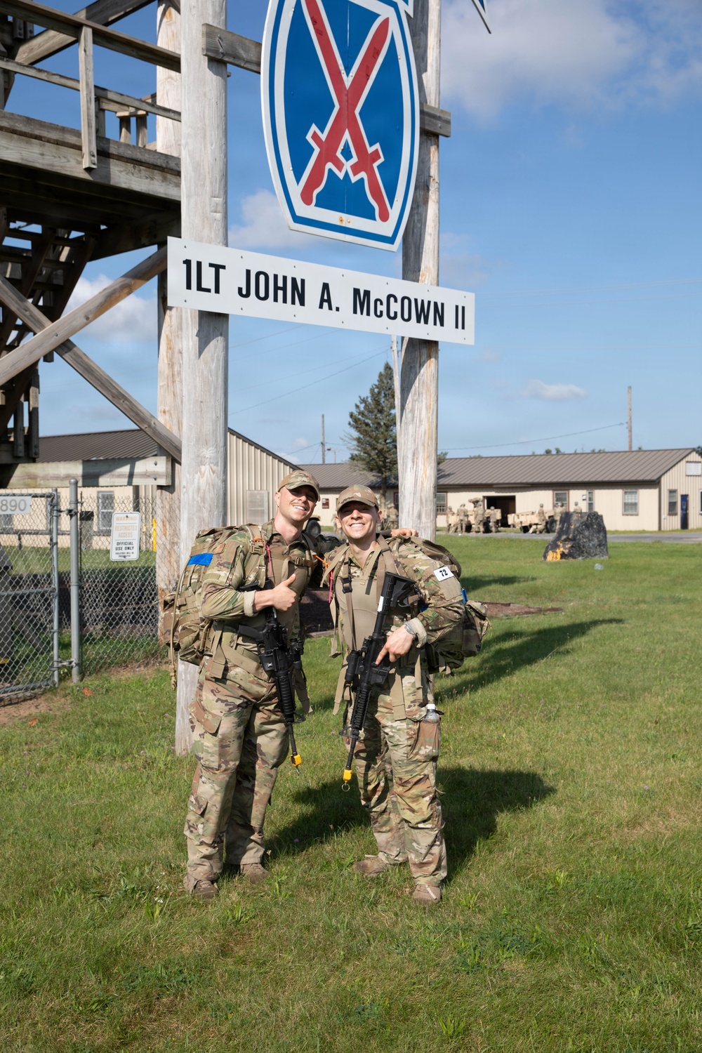 Two soldiers in full combat gear, including rucksacks and rifles, pose smiling beneath a sign reading '1LT JOHN A. McCOWN II' and the 10th Mountain Division insignia at what appears to be Fort Drum, New York. Both are wearing MultiCam (OCP) uniforms and appear to be participating in a ruck march or military competition event, evidenced by numbered bibs and colored tape markers on their gear. The setting is a military installation with barracks and military vehicles visible in the background.