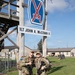 Two soldiers in full combat gear, including rucksacks and rifles, pose smiling beneath a sign reading '1LT JOHN A. McCOWN II' and the 10th Mountain Division insignia at what appears to be Fort Drum, New York. Both are wearing MultiCam (OCP) uniforms and appear to be participating in a ruck march or military competition event, evidenced by numbered bibs and colored tape markers on their gear. The setting is a military installation with barracks and military vehicles visible in the background.