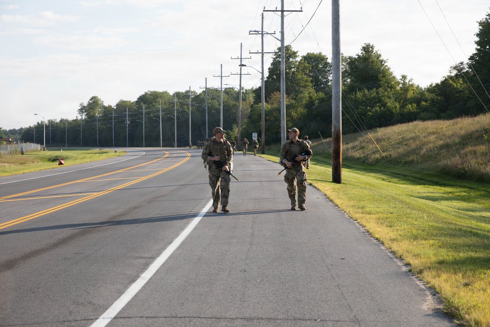 Two soldiers in camouflage uniforms and tactical gear are walking along the shoulder of a two-lane road, carrying rifles. Additional personnel are visible further down the road in the background. The setting appears to be a military installation or training area during daylight hours.