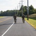 Two soldiers in camouflage uniforms and tactical gear are walking along the shoulder of a two-lane road, carrying rifles. Additional personnel are visible further down the road in the background. The setting appears to be a military installation or training area during daylight hours.