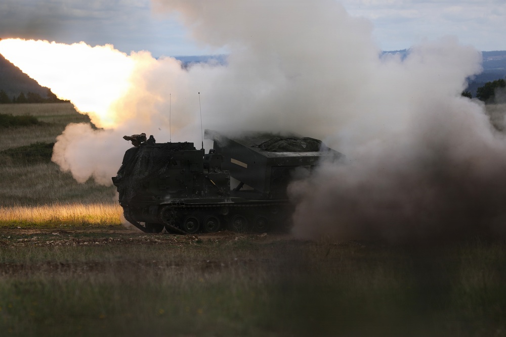 A tracked military multiple launch rocket system (MLRS) is captured mid-fire, with a rocket visibly launching and producing a large plume of white smoke and bright flame. The vehicle appears to be operating in an open field or training range during low-light conditions, likely dusk or dawn. The image documents an active rocket artillery firing event.