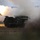 A tracked military multiple launch rocket system (MLRS) is captured mid-fire, with a rocket visibly launching and producing a large plume of white smoke and bright flame. The vehicle appears to be operating in an open field or training range during low-light conditions, likely dusk or dawn. The image documents an active rocket artillery firing event.