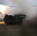 A tracked military multiple launch rocket system (MLRS) is captured mid-fire, with a rocket visibly launching and producing a large plume of white smoke and bright flame. The vehicle appears to be operating in an open field or training range during low-light conditions, likely dusk or dawn. The image documents an active rocket artillery firing event.