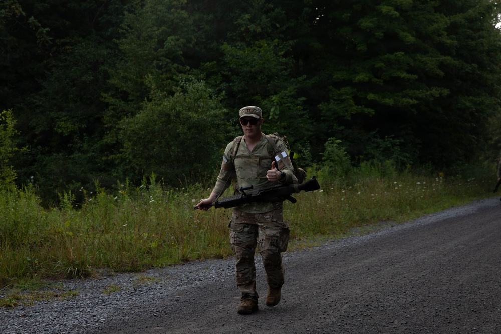 A soldier in full multicam/OCP camouflage uniform is walking along a gravel road through a wooded area. The individual is carrying what appears to be an M4/M16-style rifle and wearing a rucksack, suggesting a ruck march or field training exercise. The soldier is giving a thumbs-up gesture toward the camera.