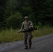 A soldier in full multicam/OCP camouflage uniform is walking along a gravel road through a wooded area. The individual is carrying what appears to be an M4/M16-style rifle and wearing a rucksack, suggesting a ruck march or field training exercise. The soldier is giving a thumbs-up gesture toward the camera.