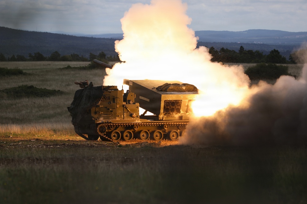 A tracked military multiple launch rocket system (MLRS) is captured mid-fire, with a rocket visibly launching from the elevated launcher pod accompanied by intense flame and large plumes of white smoke. The vehicle appears to be operating in an open field or training range during low-light conditions, likely dusk or dawn. The surrounding terrain features grassland, tree lines, and distant hills.