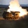 A tracked military multiple launch rocket system (MLRS) is captured mid-fire, with a rocket visibly launching from the elevated launcher pod accompanied by intense flame and large plumes of white smoke. The vehicle appears to be operating in an open field or training range during low-light conditions, likely dusk or dawn. The surrounding terrain features grassland, tree lines, and distant hills.