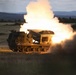 A tracked military multiple launch rocket system (MLRS) is captured mid-fire, with a rocket visibly launching from the elevated launcher pod accompanied by intense flame and large plumes of white smoke. The vehicle appears to be operating in an open field or training range during low-light conditions, likely dusk or dawn. The surrounding terrain features grassland, tree lines, and distant hills.