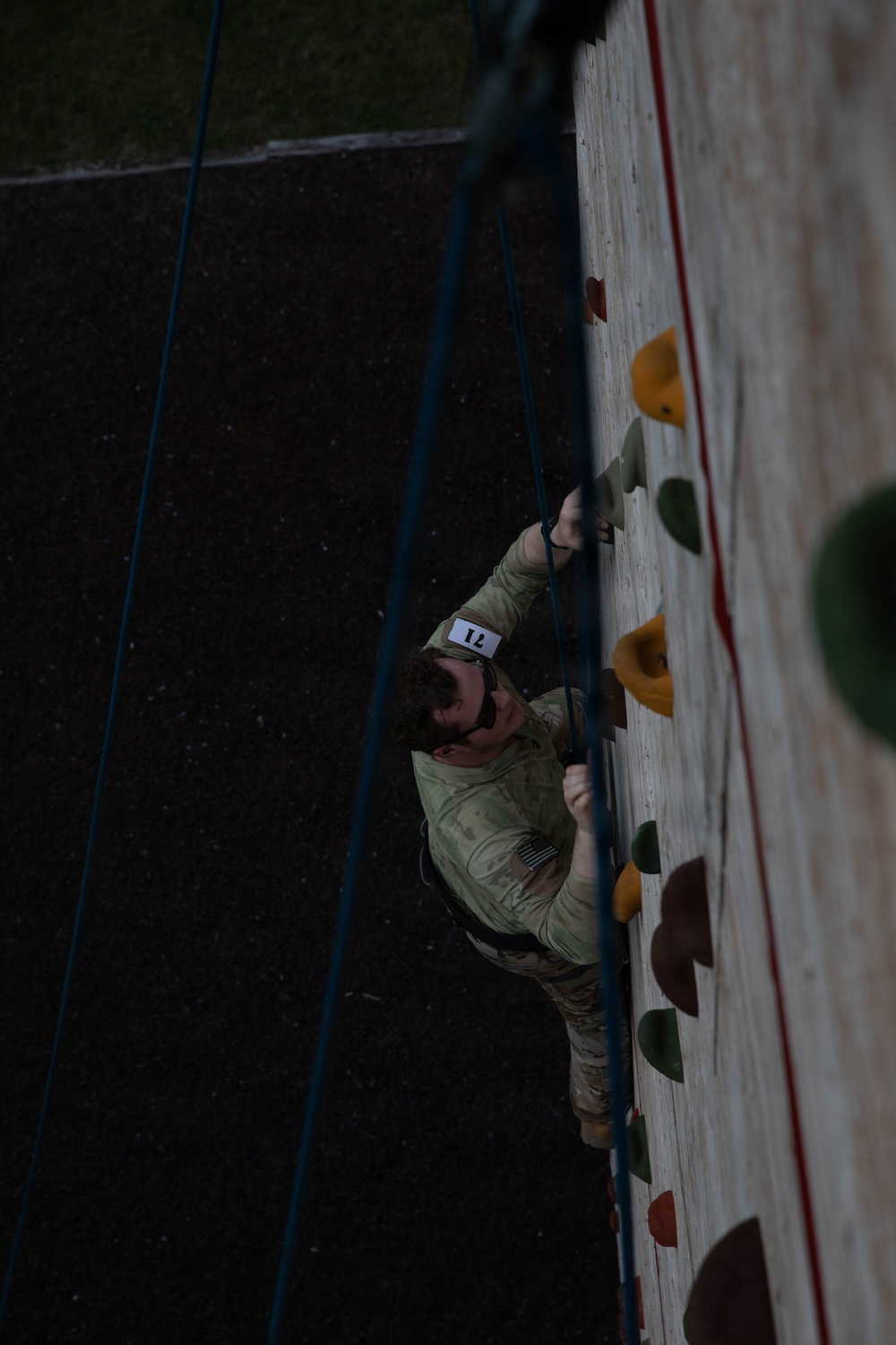 A person in military camouflage uniform is climbing an outdoor climbing wall, photographed from above. The climber wears a numbered bib (number 17, appearing reversed due to angle) and a safety harness connected to blue belay ropes. The image is taken from a top-down perspective, showing the climber mid-ascent on a wall fitted with colored climbing holds.