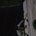 A person in military camouflage uniform is climbing an outdoor climbing wall, photographed from above. The climber wears a numbered bib (number 17, appearing reversed due to angle) and a safety harness connected to blue belay ropes. The image is taken from a top-down perspective, showing the climber mid-ascent on a wall fitted with colored climbing holds.