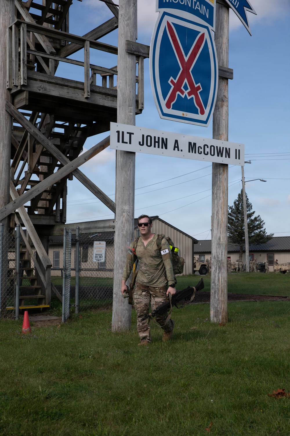 A soldier in camouflage uniform and sunglasses walks beneath a 10th Mountain Division sign at a military installation. The sign reads '1LT JOHN A. McCOWN II' and displays the 10th Mountain Division insignia with crossed bayonets. The soldier is wearing a race bib numbered '71' and carrying gear, suggesting participation in a ruck march or military fitness event.