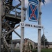 A soldier in camouflage uniform and sunglasses walks beneath a 10th Mountain Division sign at a military installation. The sign reads '1LT JOHN A. McCOWN II' and displays the 10th Mountain Division insignia with crossed bayonets. The soldier is wearing a race bib numbered '71' and carrying gear, suggesting participation in a ruck march or military fitness event.