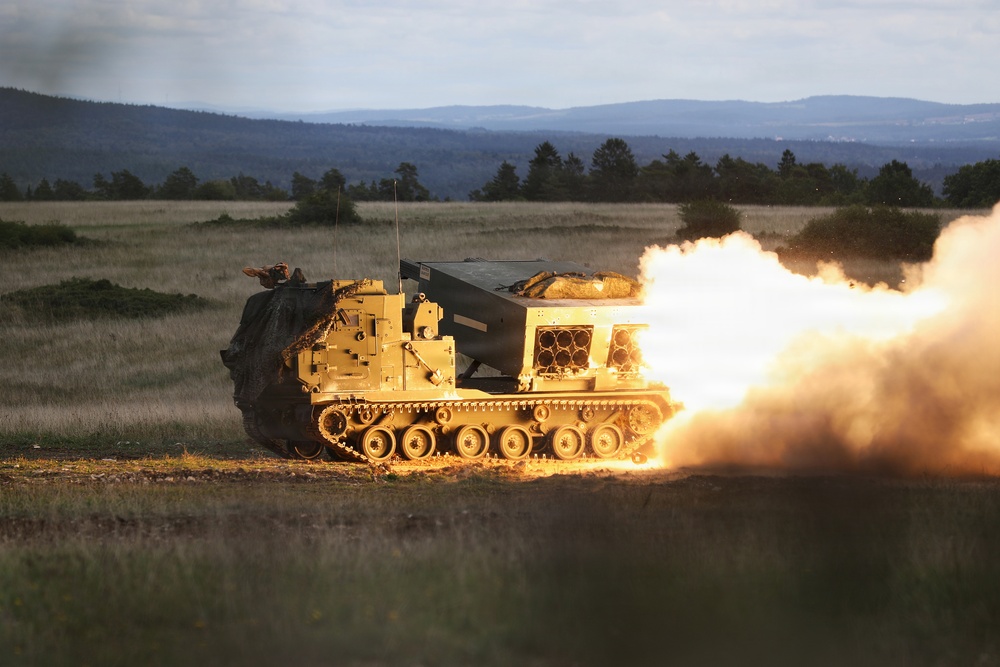 A tracked military multiple launch rocket system (MLRS), identifiable as an M270 or similar platform, is captured mid-fire in an open field during low-light conditions. A rocket or missile is visibly launching from the pod launcher, producing a large plume of fire, smoke, and exhaust to the right side of the vehicle. The setting appears to be a military training range or exercise area, with forested hills and open grassland in the background.