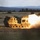 A tracked military multiple launch rocket system (MLRS), identifiable as an M270 or similar platform, is captured mid-fire in an open field during low-light conditions. A rocket or missile is visibly launching from the pod launcher, producing a large plume of fire, smoke, and exhaust to the right side of the vehicle. The setting appears to be a military training range or exercise area, with forested hills and open grassland in the background.