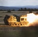 A tracked military multiple launch rocket system (MLRS), identifiable as an M270 or similar platform, is captured mid-fire in an open field during low-light conditions. A rocket or missile is visibly launching from the pod launcher, producing a large plume of fire, smoke, and exhaust to the right side of the vehicle. The setting appears to be a military training range or exercise area, with forested hills and open grassland in the background.