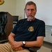 A middle-aged man with gray-streaked hair and a beard sits in a classroom or briefing room with arms crossed, wearing a dark navy t-shirt bearing a military logistics unit emblem. The setting appears to be a military facility, evidenced by a unit patch/insignia visible on the wall in the background. His posture and expression convey a serious, composed demeanor.