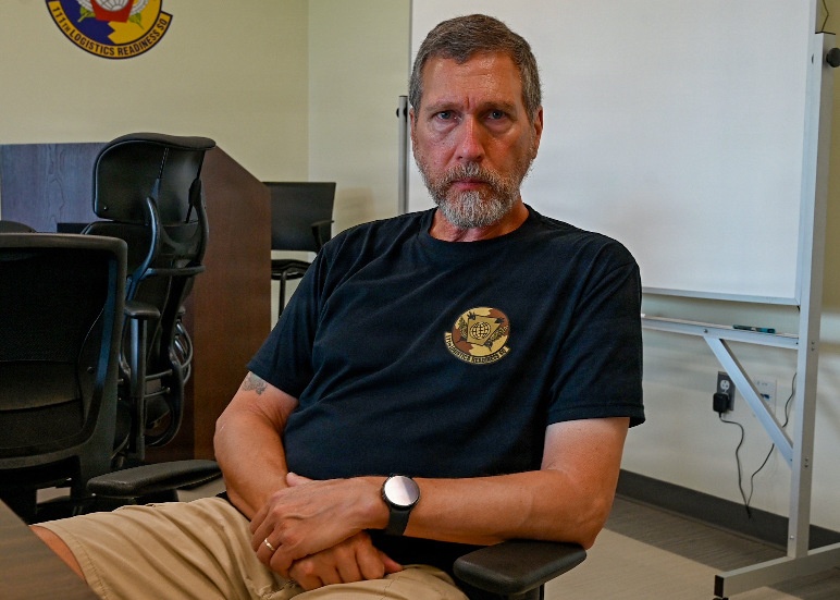 A middle-aged man with gray-streaked hair and a beard sits in a classroom or briefing room with arms crossed, wearing a dark navy t-shirt bearing a military logistics unit emblem. The setting appears to be a military facility, evidenced by a unit patch/insignia visible on the wall in the background. His posture and expression convey a serious, composed demeanor.