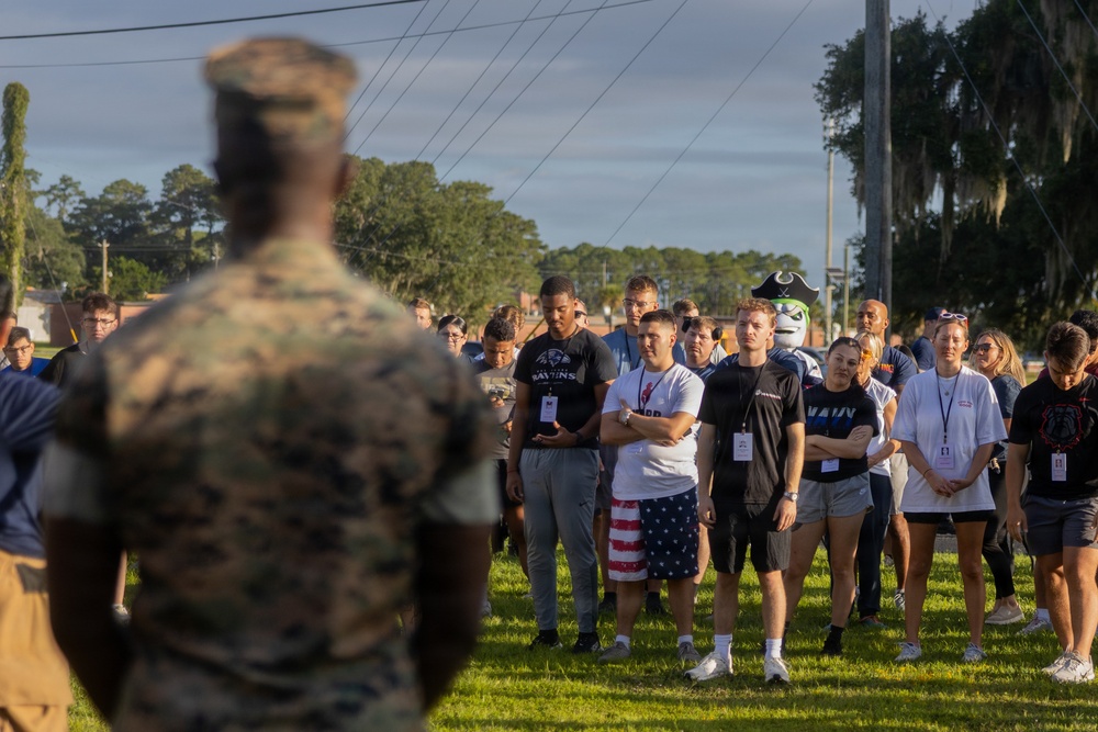 A U.S. Marine in camouflage uniform addresses a large group of civilians on a grassy field, likely at a military base. The civilians, wearing casual clothing and lanyards/badges, appear to be participants in an orientation or community outreach event. A sports team mascot wearing a pirate-style costume is visible among the crowd in the background.