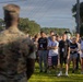 A U.S. Marine in camouflage uniform addresses a large group of civilians on a grassy field, likely at a military base. The civilians, wearing casual clothing and lanyards/badges, appear to be participants in an orientation or community outreach event. A sports team mascot wearing a pirate-style costume is visible among the crowd in the background.