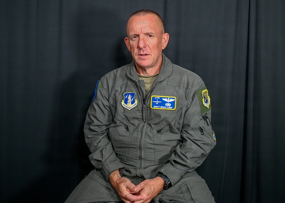 A male military personnel member wearing a gray flight suit is seated against a dark curtain backdrop, appearing to be in an interview or recording setting. His name tag reads 'SMSGT BRIAN CURRY' with a callsign 'HUBBY', and he wears Air National Guard insignia patches. He is mid-speech with hands clasped in his lap.