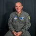 A male military personnel member wearing a gray flight suit is seated against a dark curtain backdrop, appearing to be in an interview or recording setting. His name tag reads 'SMSGT BRIAN CURRY' with a callsign 'HUBBY', and he wears Air National Guard insignia patches. He is mid-speech with hands clasped in his lap.