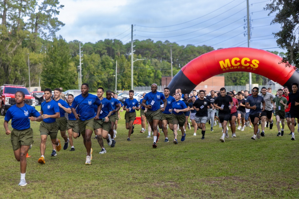 A large group of military personnel and civilians are participating in a running event on a grass field, starting from beneath an inflatable arch branded 'MCCS Semper Fit.' The front runners are wearing matching blue t-shirts and olive/khaki shorts, consistent with Marine Corps athletic uniforms. The event appears to be an organized fitness run or fun run held on a military installation.