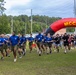 A large group of military personnel and civilians are participating in a running event on a grass field, starting from beneath an inflatable arch branded 'MCCS Semper Fit.' The front runners are wearing matching blue t-shirts and olive/khaki shorts, consistent with Marine Corps athletic uniforms. The event appears to be an organized fitness run or fun run held on a military installation.