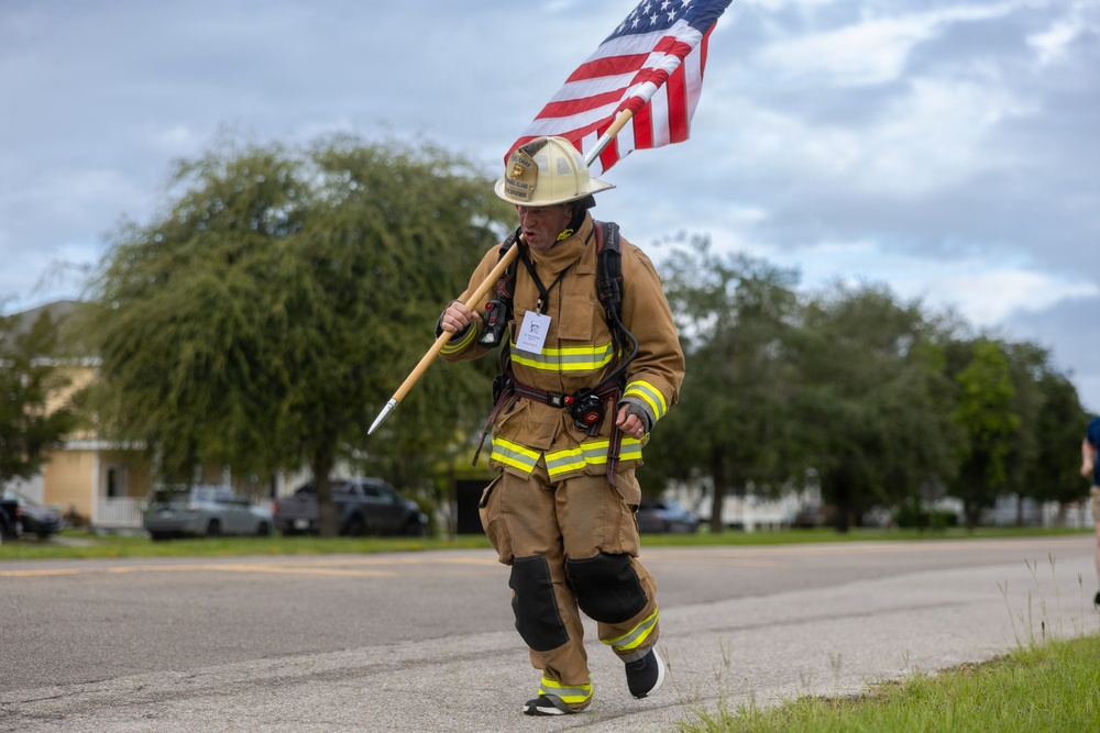 A firefighter in full turnout gear is running along a road while carrying an American flag on a wooden pole. The individual is wearing a white helmet, tan/yellow reflective bunker gear, knee pads, and appears to have a breathing apparatus or pack on their back. This appears to be a memorial run or tribute event, likely honoring fallen first responders.