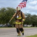 A firefighter in full turnout gear is running along a road while carrying an American flag on a wooden pole. The individual is wearing a white helmet, tan/yellow reflective bunker gear, knee pads, and appears to have a breathing apparatus or pack on their back. This appears to be a memorial run or tribute event, likely honoring fallen first responders.