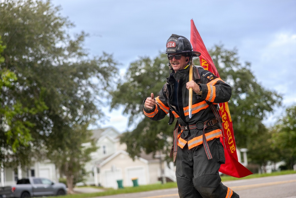 A firefighter in full turnout gear walks along a residential street while carrying a red 'Fire Rescue' flag on a wooden pole. The individual is wearing a helmet numbered '524' with 'Fire Rescue' text and is giving a thumbs-up gesture toward the camera. The setting appears to be a suburban neighborhood with houses and trees visible in the background.