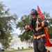 A firefighter in full turnout gear walks along a residential street while carrying a red 'Fire Rescue' flag on a wooden pole. The individual is wearing a helmet numbered '524' with 'Fire Rescue' text and is giving a thumbs-up gesture toward the camera. The setting appears to be a suburban neighborhood with houses and trees visible in the background.